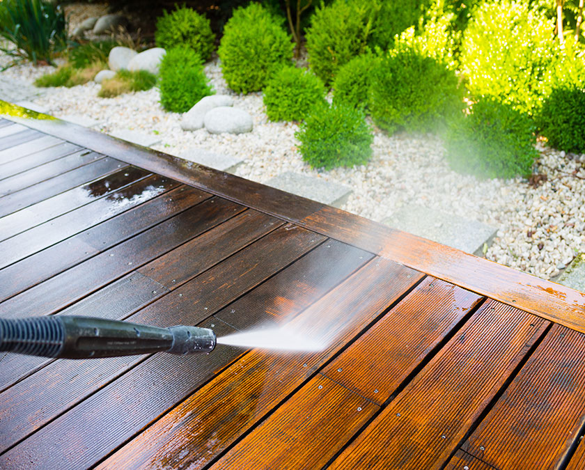 Power wash cleaning of wooden deck, with white sand and green shrubs in the background.