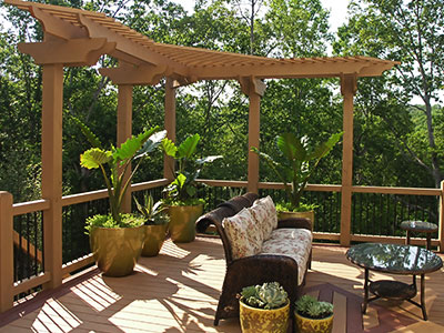 Wooden deck with black metal railing, wooden shade cover, rattan couch, glass table, and trees in the background.